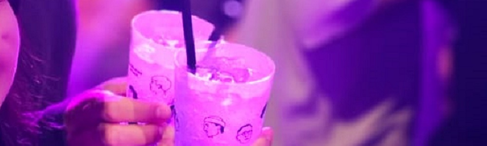 Zwei Plastikbecher mit Strohhalm und Getränk darin, werden von zwei Händen, in einer Menschenmenge, gehalten. Die Szene ist in diffuses rosa Licht getaucht.