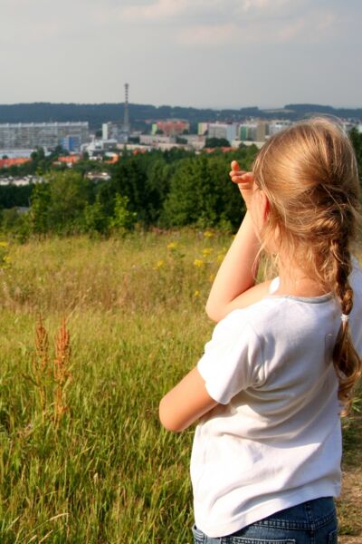 Ein Mädchen ist von Hinten zu sehen, es schaut in eine Landschaft. Vorne Felder und Bäume, dahinter ein Stadt.