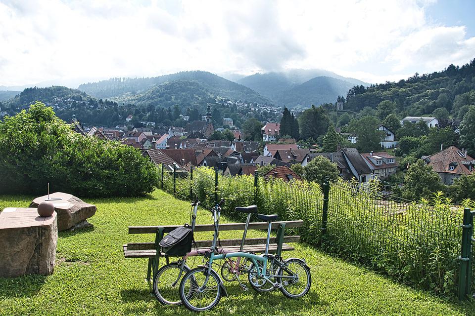 Die Abbildung zeigt den Blick von oben auf ein Dorf. Im Vordergrund des Bildes ist ein Rastplatz mit einer Bank zu sehen, an die zwei Fahrräder angelehnt sind.