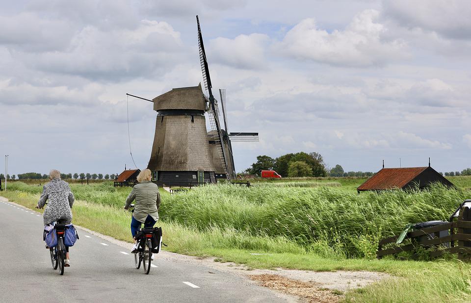 Die Abbildung zeigt zwei Fahrradfahrerinnen auf einer Straße. Im Hintergrund sind zwei alte Mühlen zu sehen.