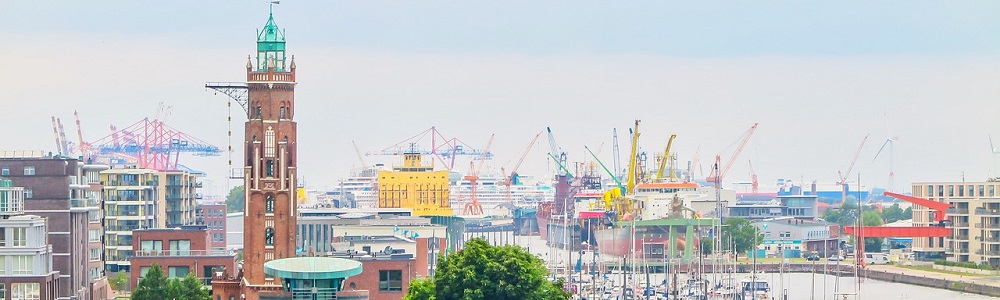 Eine Aufnahme aus höherer Höhe von Bremerhaven. Zu sehen sind ein Turm, Häuser, Straßen, Schiffe, Kräne und einem Krezfahrtschiff im Hintergurnd und das Havenbecken mit Segelschiffen.